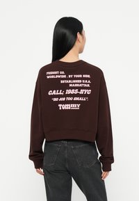 Felpa corta marrone con testo rosa sul retro. Il testo include "FREIGHT CO.", "CALL; 1985-NYC" e "TOMMY jeans". Tessuto morbido, vestibilità rilassata.