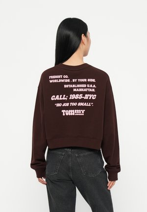 Bruine cropped sweatshirts met roze tekst op de achterkant. De tekst bevat "FREIGHT CO.", "BEL; 1985-NYC" en "TOMMY jeans". Zachte stof, relaxte pasvorm.