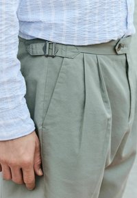 Jasnozielone, plisowane spodnie wykonane z teksturowanej tkaniny, z czarną klamrą i bocznymi kieszeniami, sparowane z niebieską i białą koszulą w kratę.