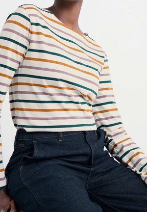 Personne portant une chemise blanche à manches longues avec des rayures vertes, orange et violettes et un jean en denim bleu foncé, assise devant un fond blanc.