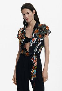 Short-sleeve, multi-patterned shirt featuring bold floral and geometric designs in vibrant colors, secured with a tied waist.