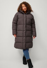 Cappotto imbottito grigio scuro con cappuccio, caratterizzato da un design trapuntato, maniche lunghe e un taglio di lunghezza media. Indossato sopra jeans blu e stivali neri.
