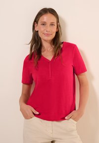 Haut rouge à manches courtes avec un décolleté en V et une texture subtile. Le mannequin pose les mains dans les poches, portant un pantalon de couleur claire.