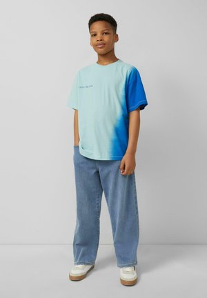 Garçon debout devant un fond uni, portant un t-shirt tie-dye bleu clair et bleu avec texte, jean large et baskets blanches.