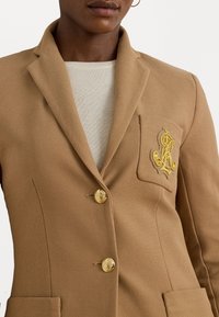 Svetle hnědý blazer s texturovanou tkaninou, dvěma zlatými knoflíky a přední kapsou s komplikovaným zlatým emblémem. Klasický design límečku.