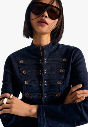 Kvinde med kort mørkt hår iført store mørke solbriller og en mørkeblå jakke med dekorative knapper, krydser armene.