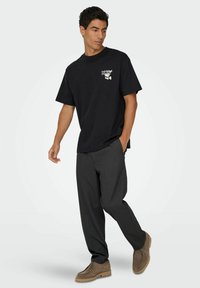Schwarzes Baumwoll-T-Shirt mit kurzen Ärmeln und Grafik auf der Brust, kombiniert mit grauen Hosen und braunen Wildlederschuhen.