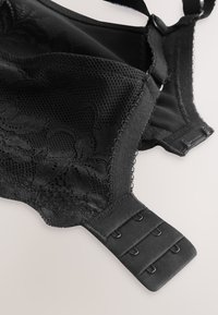 Gros plan sur un soutien-gorge en dentelle noire montrant un motif floral, un tissu en maille, des bretelles réglables et une fermeture dos à trois crochets sur un fond clair.
