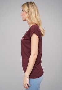 Burgunderfarget kortermet t-skjorte med avslappet passform og rund hals, med hvit grafisk design på forsiden. Brukt sammen med lyseblå jeans.