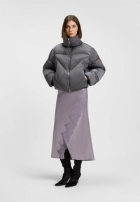 Grå pufferjacka med hög krage och dragkedja, bärs över en lilafärgad satenskirt med en rufsig spetsdetalj. Långa svarta stövlar fulländar outfiten.