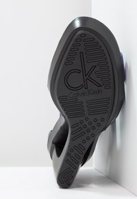 Semelle de chaussure noire avec motifs de grip texturés, présentant un logo "ck" en relief et un design lisse et courbé. Accents en matériau bleu foncé.
