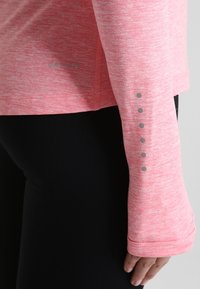 Jasnoróżowa, długosleeveowa koszulka sportowa z textured tkaniny, z odblaskowymi szarymi kropkami na rękawach oraz subtelnym logo na końcu.