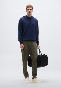 Tengerészkék kapucnis pulóver, olívazöld oldalsávos melegítőnadrág, krémszínű sportcipők. A modell egy fekete utazótáskát tart. Egyszerű, hétköznapi öltözék.