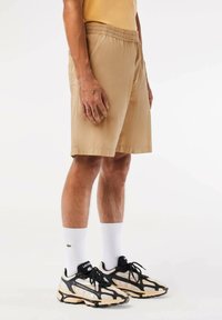 Pantalones cortos beige con cinturilla elástica, bolsillos laterales y un corte relajado. Llevados con calcetines blancos y zapatillas deportivas negras y crema.