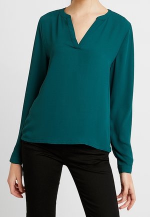 Kobieta ubrana w bluzkę w kolorze teal z długim rękawem i dekoltem w kształcie litery V, do której dobrano czarne dopasowane spodnie, na jednolitym tle.