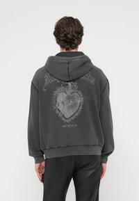 Γκρι φαρδιά φούτερ με κουκούλα, με σχέδιο γραφικής καρδιάς και το κείμενο «MCMXCIV» στην πλάτη. Κατασκευασμένο από βαμβάκι, με ρυθμιζόμενο κορδόνι στην κουκούλα και ριμπ μανσέτες.