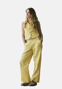 Chaleco de lino amarillo con cinco botones, combinado con pantalones amarillos de corte holgado, que presentan una textura suave y un sutil brillo.