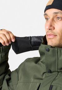 Zielona wodoodporna kurtka z czarnymi akcentami, wysoki kołnierz i regulowane mankiety. Posiada zatrzaski i teksturowany materiał dla lepszej izolacji.