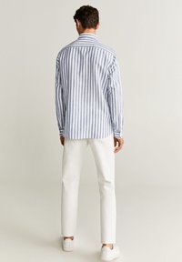 Muž oblečený v modro-bílém pruhovaném dlouhém rukávu, bílých kalhotách a bílých teniskách, stojící čelem k jednoduchému světlému pozadí.