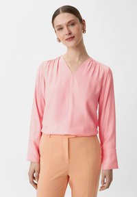 Rosa langärmlige Bluse mit V-Ausschnitt und gerafften Schultern, kombiniert mit pfirsichfarbenen Hosen. Glatte Textur, minimalistisches Design.