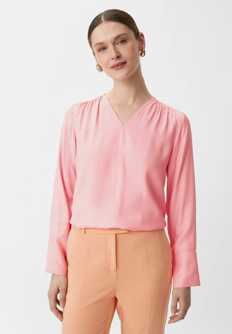 Rosa langärmlige Bluse mit V-Ausschnitt und gerafften Schultern, kombiniert mit pfirsichfarbenen Hosen. Glatte Textur, minimalistisches Design.