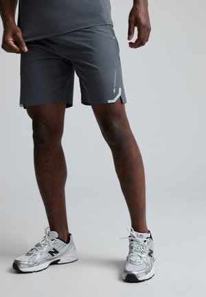 En person iført mørkegrå sportsshorts og sølvfarvede New Balance løbesko, der står mod en ensfarvet lys baggrund.