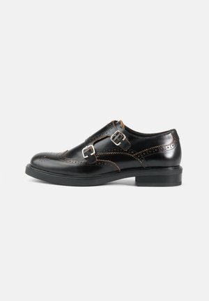 Scarpa nera in pelle con doppia fibbia monk, perforazioni brogue, fibbie argento e tacco basso su sfondo bianco.