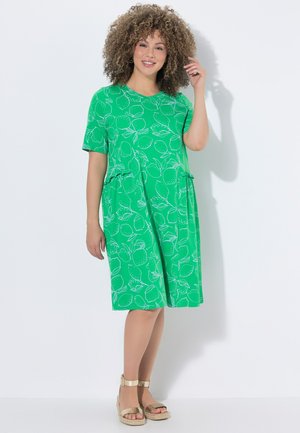 Kobieta o kręconych włosach, ubrana w zieloną sukienkę za kolano z wzorem cytryn i złote sandały na koturnie, uśmiechnięta, stojąca przy białej ścianie.