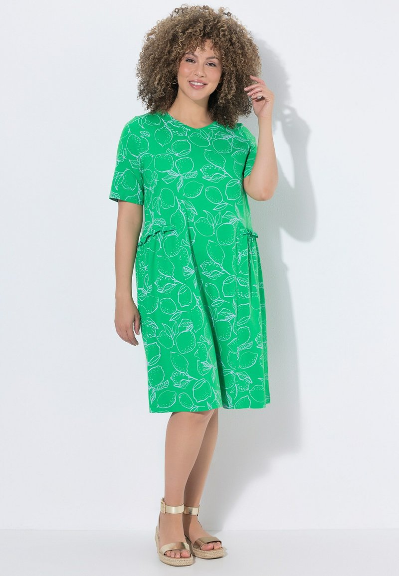 Femme aux cheveux bouclés portant une robe verte mi-longue avec un motif de citron et des sandales compensées dorées, souriante, debout contre un mur blanc.