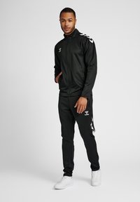 Survêtement noir en tissu lisse, composé d'une veste zippée et de pantalons fuselés ; présente des logos blancs et des accents en chevrons sur les manches.