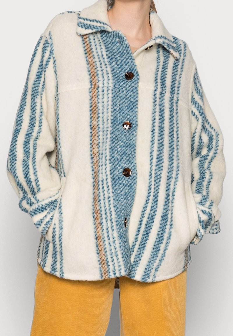 Femme portant un manteau crème boutonné avec des rayures verticales bleues et brunes et un pantalon en velours jaune, mains dans les poches.