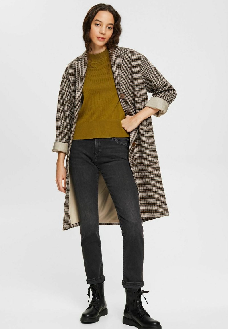 Manteau à carreaux marron et beige, superposé sur un pull côtelé moutarde, associé à un jean noir et des bottes de combat à lacets.