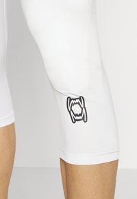 Hvide atletiske kompressionsshorts med en glat overflade, prydet med et sort logo på låret og en tætsiddende længde lige over knæet.