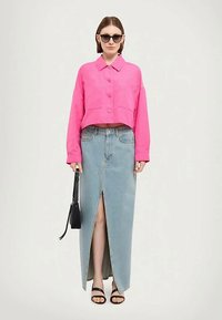 Kvinne som har på seg svarte solbriller, rosa knappet kort jakke, lyseblå denim skjørt med frontslitt, svarte sandaler, og holder en liten svart håndveske.