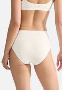 Femme portant un slip taille haute blanc et un soutien-gorge de sport assorti, vue de dos contre un fond clair.