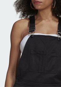 Černé kšandy s nastavitelnými popruhy, kovovými prvky a přední kapsou. Tkanina vypadá odolně a má vertikální švy.