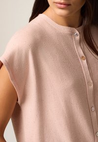 Vrouw in een lichtroze, knooploze mouwloze trui met subtiele sprankelende details, waarbij haar kin en een deel van haar lange donkere haar zichtbaar zijn.