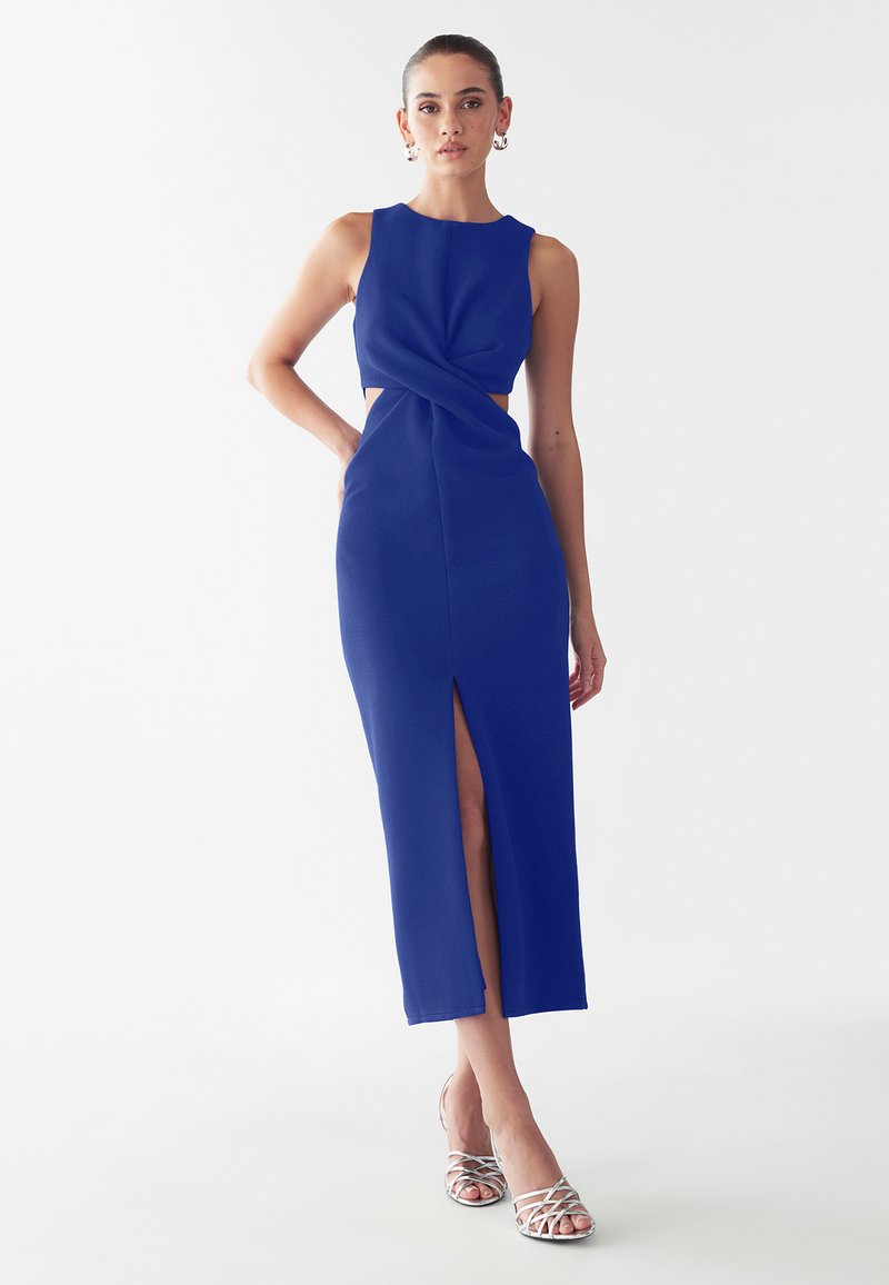 Royal blue fitted dress with a twist detail at the waist, featuring a thigh-high slit and sleeveless design. Model wears silver sandals.