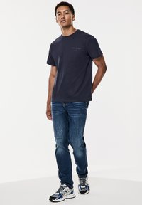 Námořnická modrá bavlněná tričko s krátkými rukávy a malým logem "Vinyl Club". Nosené s modrými džíny a barevnými sportovními botami.