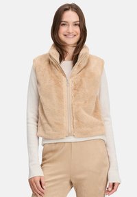 Beżowa kamizelka z futra syntetycznego z wysokim kołnierzem i zamkiem z przodu. Miękka tekstura i dopasowany krój, w połączeniu z jasnym swetrem pod spodem.