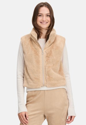 Beige neppelsvest met een hoge kraag en een ritssluiting aan de voorkant. Zachte textuur en aansluitend ontwerp, gecombineerd met een lichtgekleurde trui eronder.