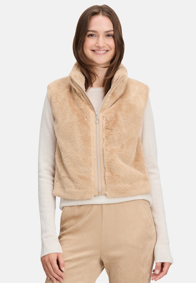 Beżowa kamizelka z futra syntetycznego z wysokim kołnierzem i zamkiem z przodu. Miękka tekstura i dopasowany krój, w połączeniu z jasnym swetrem pod spodem.