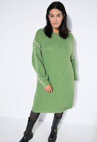 Grünes gestricktes Kleid mit langen Ärmeln, das eine strukturierte Verarbeitung und kontrastierende weiße Seitennaht aufweist, kombiniert mit schwarzen Stiefeln.