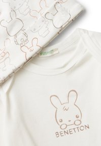 Bílý dětský body a kalhoty s hnědými kreslenými králičími obličeji a textem "Benetton" na přední straně body.
