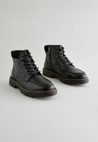 Botas de cuero negro hasta el tobillo con cordones y cremallera lateral, acabado texturizado, puntera redonda, suela de goma y detalle de costura en marrón contrastante.