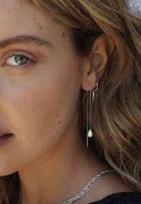 Silverörhängen med en böjd ring och hängande kedjor med en droppformad pärla och en spetsig detalj. Texturerad hud och vågigt hår.