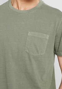 Homme portant un t-shirt uni vert olive à col rond avec une poche sur le côté gauche de la poitrine, montré de la poitrine au cou.
