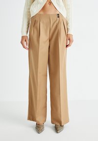 Pantalons larges beiges avec plis avant, deux boutons foncés à la taille et texture lisse. Associés à des chaussures argentées à nez pointu.
