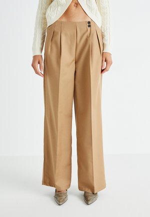 Pantalons larges beiges avec plis avant, deux boutons foncés à la taille et texture lisse. Associés à des chaussures argentées à nez pointu.