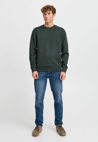 Mørkegrønn sweatshirt med rund hals, lange ermer og en glatt tekstur, sammen med blå jeans og beige sko.